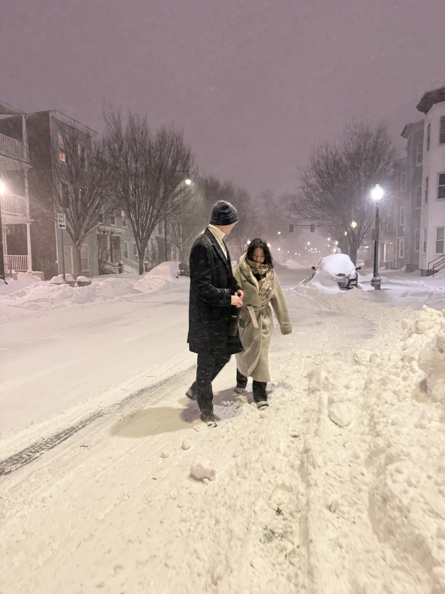 Two figures walking arm in arm down an unploughed street through heavy snow at night