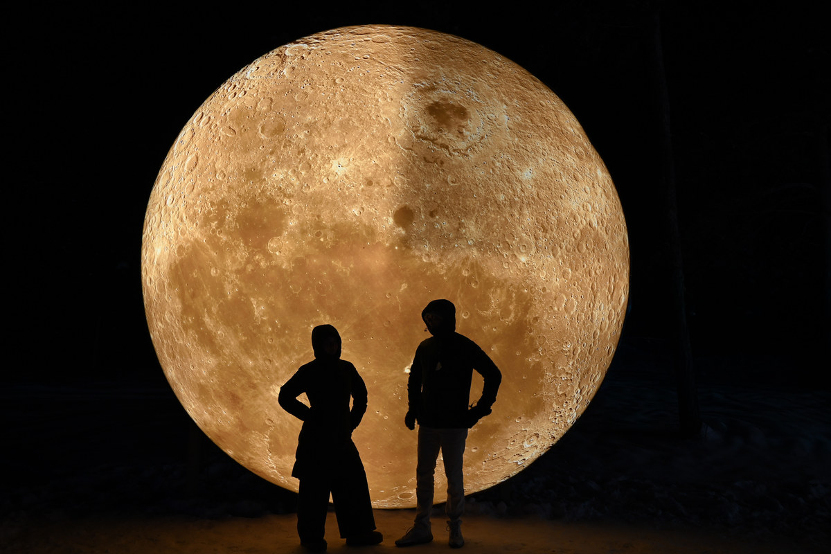 Two silhouetted figures in front of a vast illuminated model of the moon