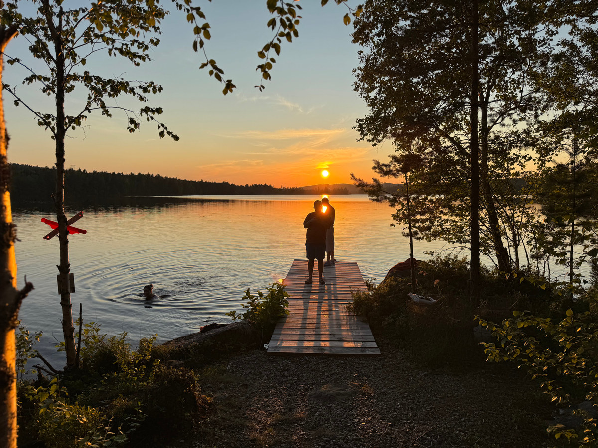Two figures silhouetted on a wooden jetty as the sun sets over a Swedish lake