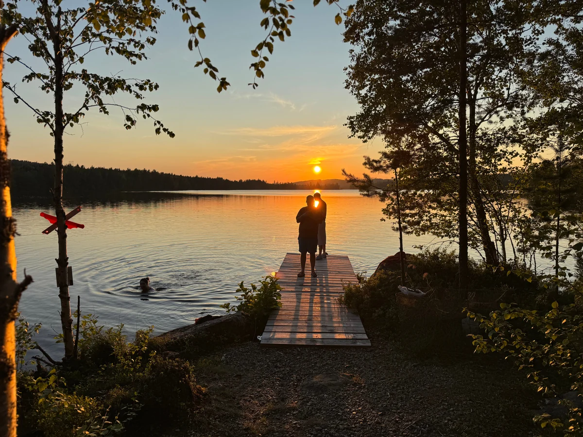 Two figures silhouetted on a wooden jetty as the sun sets over a Swedish lake